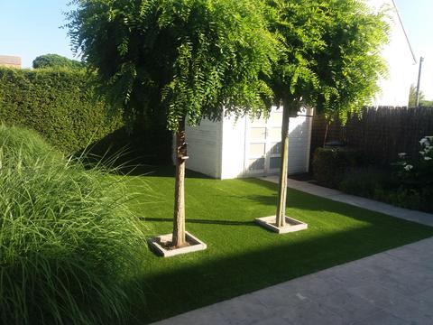 Tuin in Brugge met kunstgras rond twee bolacacia’s in betonnen boomspiegels, naast een wit tuinhuis