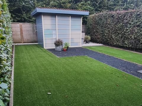 Achtertuin met kunstgras, tuinhuis en beukenhaag in Eeklo, met een goot langs het terras