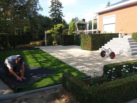 Plaatser snijdt de kunstgrasmat op maat langs de boordsteen in een tuin in Heule