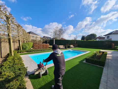 Plaatser borstelt het kunstgras op naast een zwembad tussen buxushagen, tussen Kleit en Knesselare