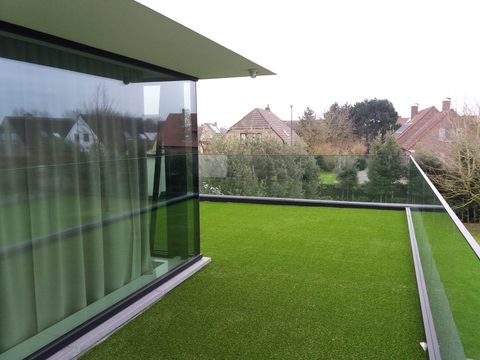 Dakterras in Maldegem met kunstgras achter een glazen balustrade, met zicht op de buurt