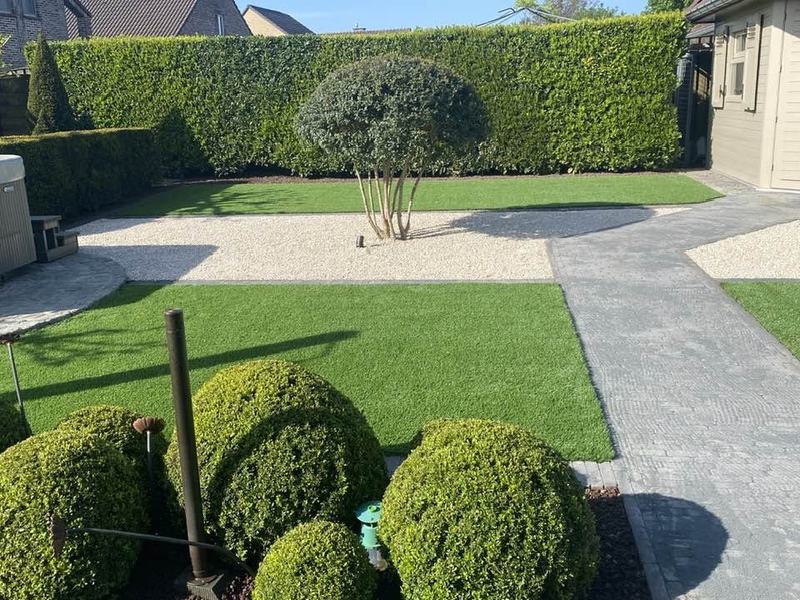 Strakke tuin in Limburg met kunstgras tussen buxusbollen, een grindvlak en een geschoren haag, gelegd door Tuinwerken Reumers