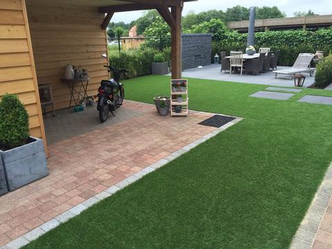 Achtertuin met kunstgras naast een overdekt terras met tuinhuis en een terras met tuinmeubelen in Limburg