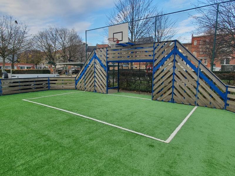 Hetzelfde buurtvoetbalveldje met een nieuwe ingezande kunstgrasmat, witte belijning, houten boarding en basketring