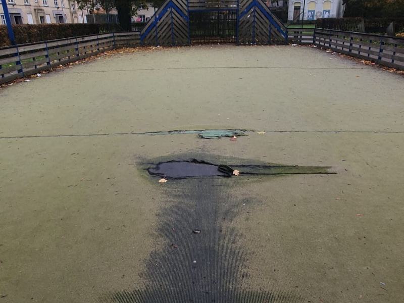 Versleten kunstgrasmat van een buurtvoetbalveldje met een groot gat, losgekomen stroken en een donker doorloopspoor voor het doel