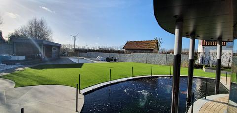 Ronde koivijver onder een zwarte luifel met daarachter een vlak kunstgras, een zwembad en een schanskorfmuur