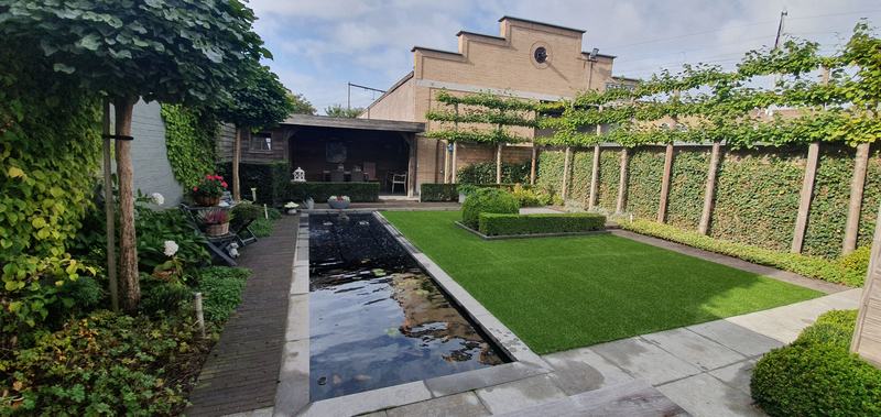 Strakke tuin met een lange rechthoekige koivijver naast een vlak kunstgras, omzoomd door leilinden en een haag