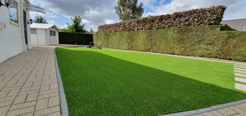 Achtertuin in Zelzate met kunstgras tussen terras, haag en tuinhuis